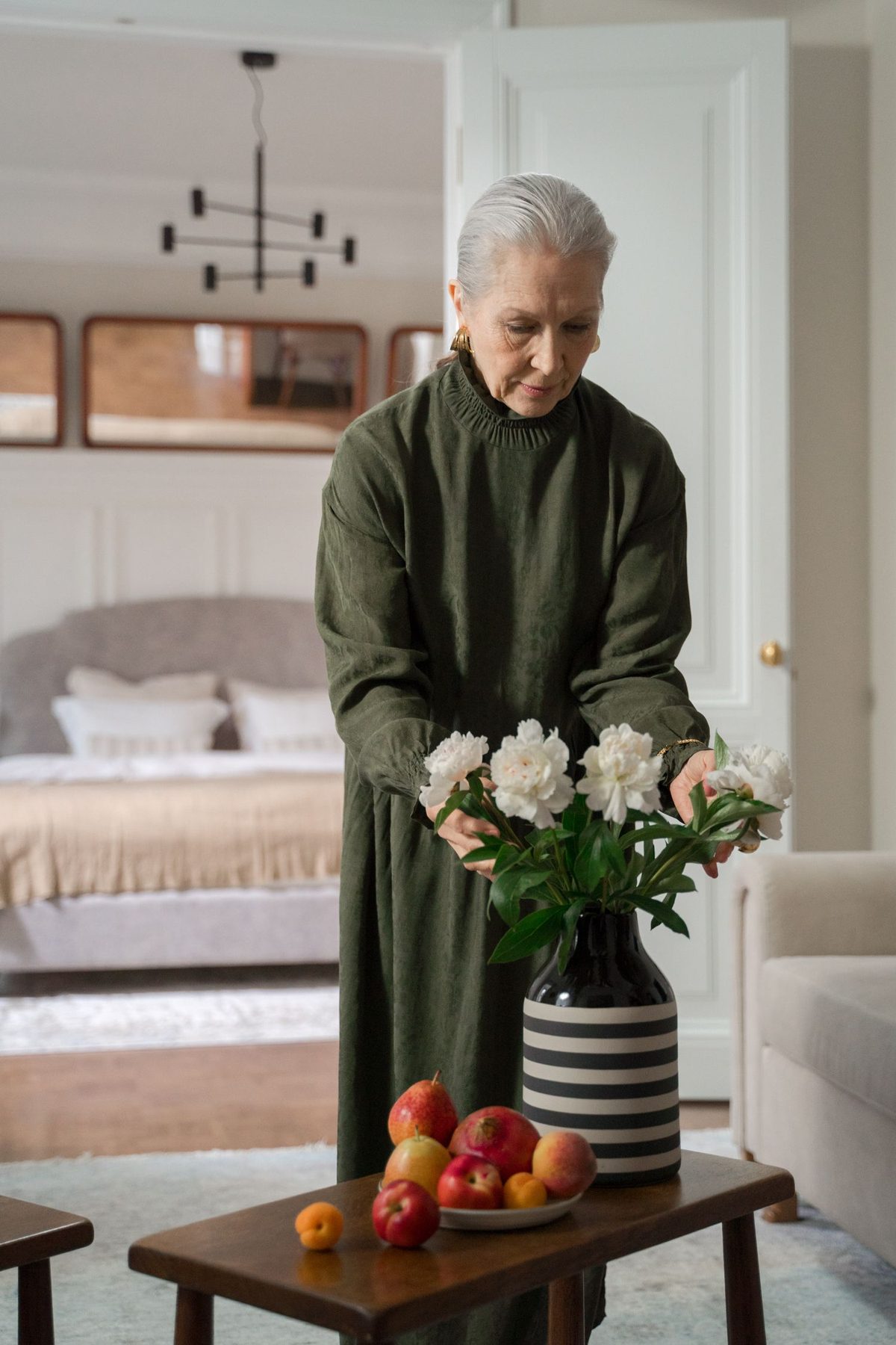 An older woman in a long green dress arranges white flowers on a side table in her own home, her bedroom visible through the doorway behind her.