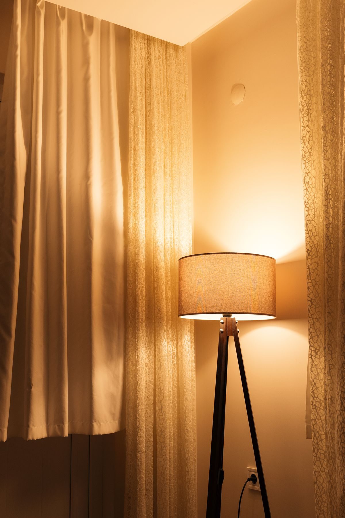 A floor lamp glowing beside drawn curtains in a quiet room at night.